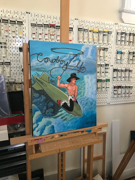'COWBOY UP' original acrylic painting on 16x20" stretched canvas. This artwork features a gay cowboy riding a falling bomb through the sky, his trailing lasso making the title: 'COWBOY UP'.  Painting shown on my easel in my art studio.